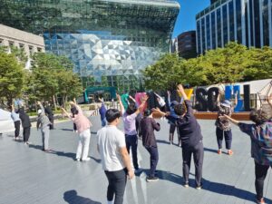 A group of older adults participating in an outdoor exercise session led by an instructor in front of the Seoul City Hall building.