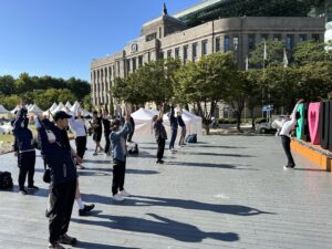 Older adults and community members participating in an outdoor group exercise session in Seoul, practicing coordinated movements in a public plaza surrounded by modern buildings.