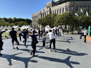 Participants of various ages performing balance and mobility exercises during an outdoor community workout in a public square in Seoul.