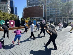 A group of older adults engaging in outdoor physical activity together in a central Seoul plaza, following guided movements under clear, sunny weather.