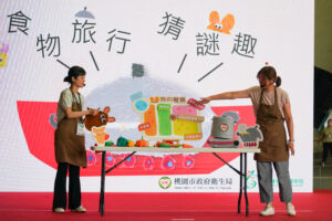 Two community nutrition educators demonstrate healthy eating concepts on stage using food models and visual boards during a public education event in Taiwan.