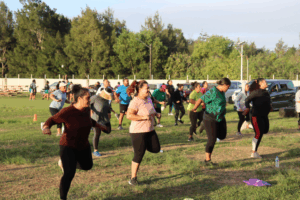 A large group of adults participating in an outdoor Zumba session on a grassy field, led by an instructor at the front.